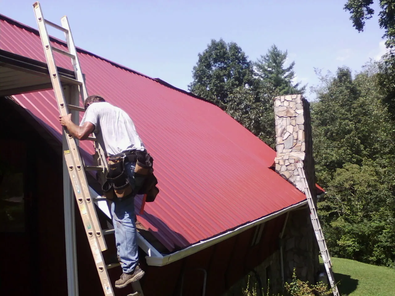 Roofer on a ladder beside a red metal roof and stone chimney — Soffit & Fascia Repair by Roofing Brothers in Harpswell